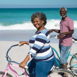 Deux séniors sur la plage avec la mère en arrière-plan tenant leur vélo sourient en direction de la caméra