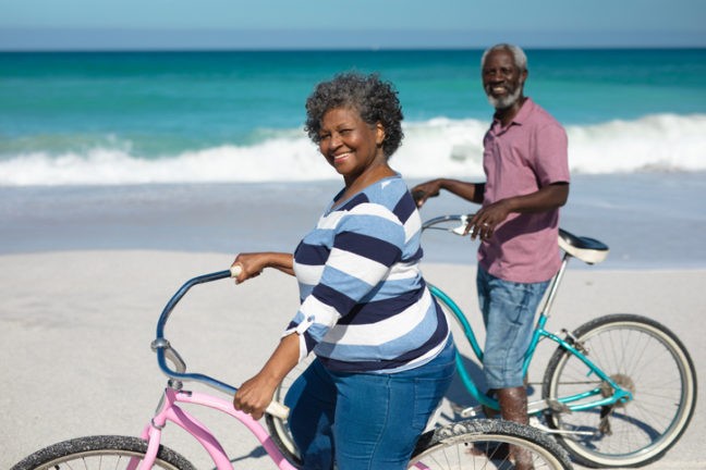 Deux séniors sur la plage avec la mère en arrière-plan tenant leur vélo sourient en direction de la caméra