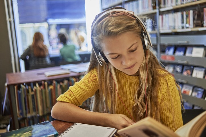 Une jeune fille avec des écouteur, les cheveux longs blonds et un pull jaune lit un livre dans une bibliothèque